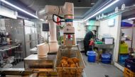 In this photo taken on June 13, 2023, a robot fries chicken as an employee washes dishes at a Robert Chicken restaurant in Seoul. (Photo by Anthony Wallace / AFP)