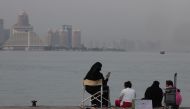 A family sits on the pavement by the Gulf waters on Doha's beachfront with the Qatari capital's landmark hotels and highrises in the background on August 6, 2024. (Photo by KARIM JAAFAR / AFP)