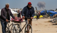 Two elderly men walk pushing bicycles while behind a cloud of smoke erupts from Israeli bombardment on the northern Gaza Strip, in Beit Lahia on March 25, 2025. Photo by Bashar TALEB / AFP