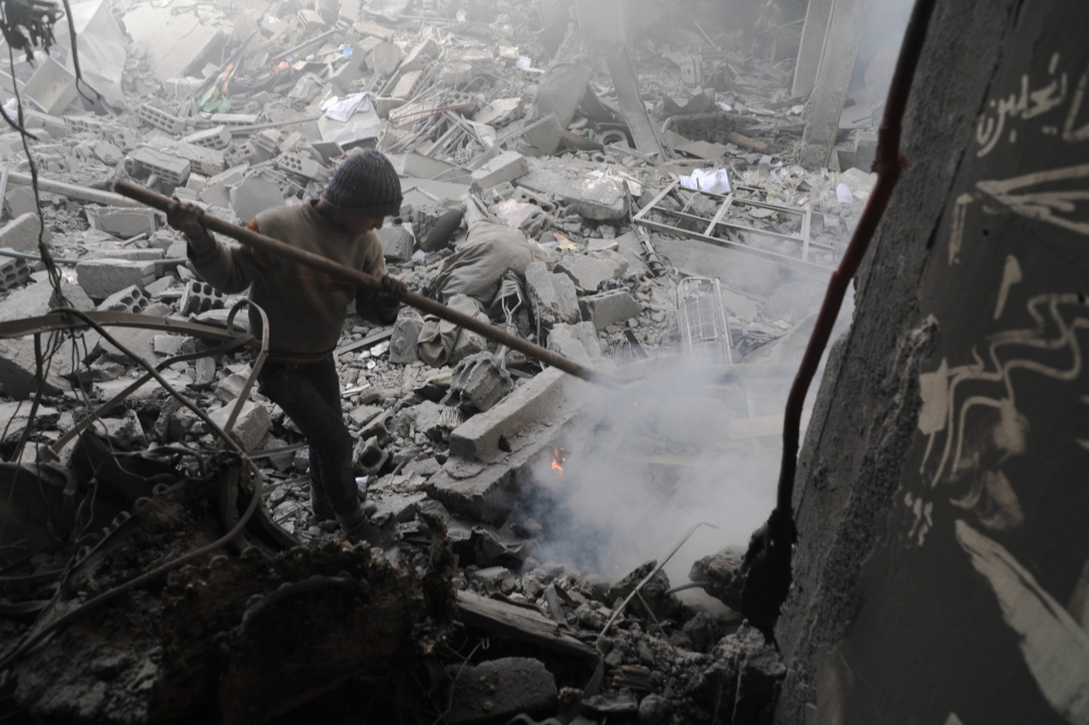 A Syrian boy puts off a fire amid destroyed buildings following government air strikes in the Eastern Ghouta rebel-held enclave of Douma, on the outskirts of the capital Damascus on March 19, 2018.   AFP / HAMZA AL-AJWEH
