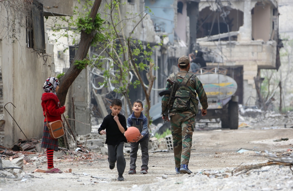 Syrian boys play with a basketball as a government soldier walks by down a street in the town of Hazzeh in Eastern Ghouta, on the outskirts of the Syrian capital Damascus, on March 28, 2018. / AFP / STRINGER