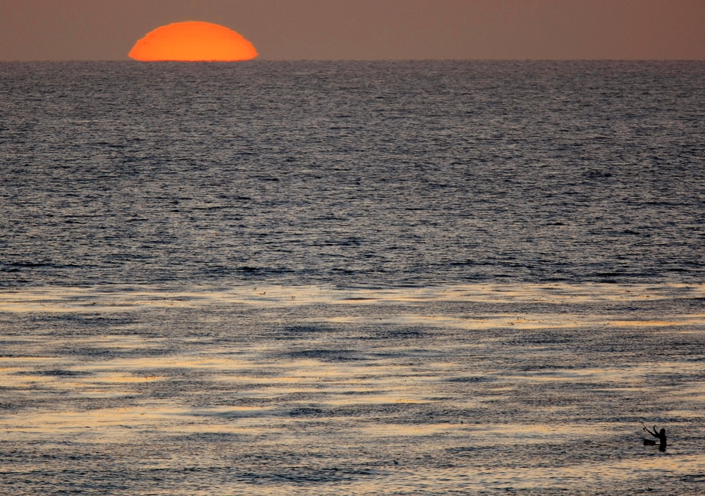 The sun sets as a fisherman casts his line in the Pacific Ocean while sitting on a surfboard off the coast of Cardiff, California April 1, 2008. REUTERS/Mike Blake/File Photo