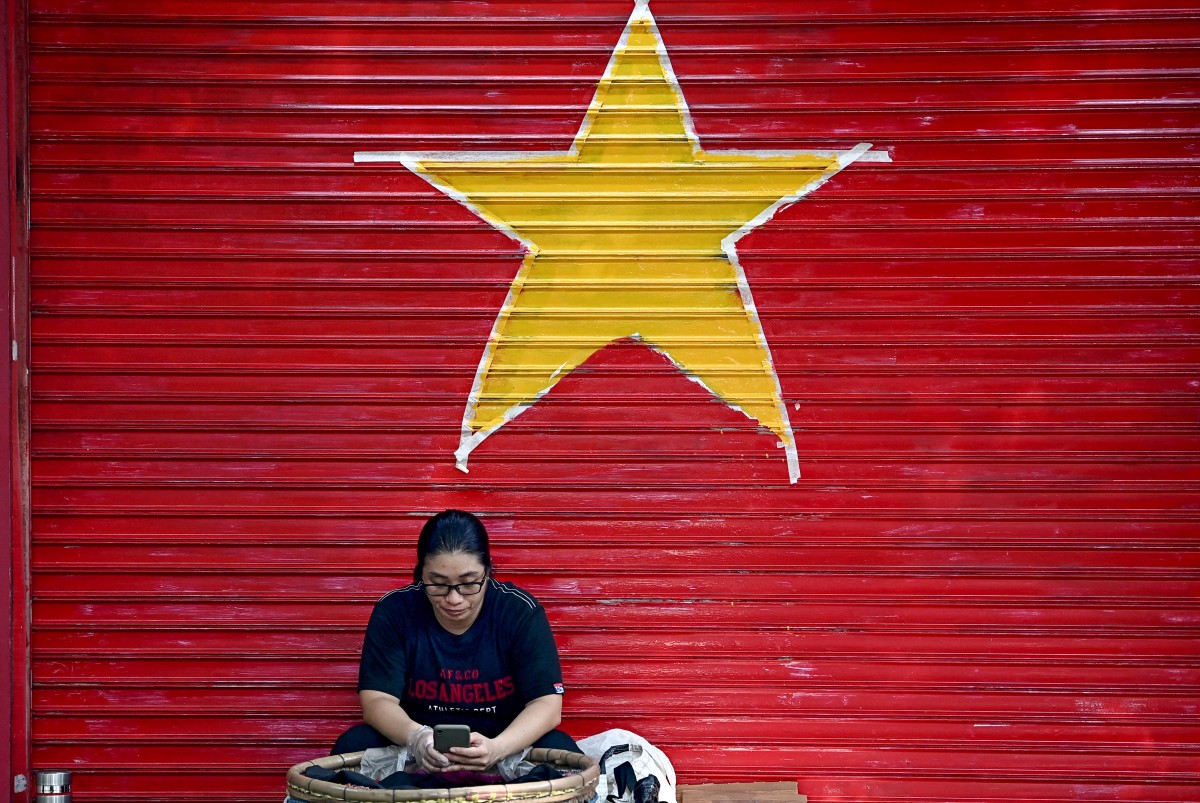 A street vendor uses her mobile phone as she sits in front of a garage door in the colours and shapes of the Vietnamese flag in Hanoi on August 28, 2024. (Photo by Nhac NGUYEN / AFP)
