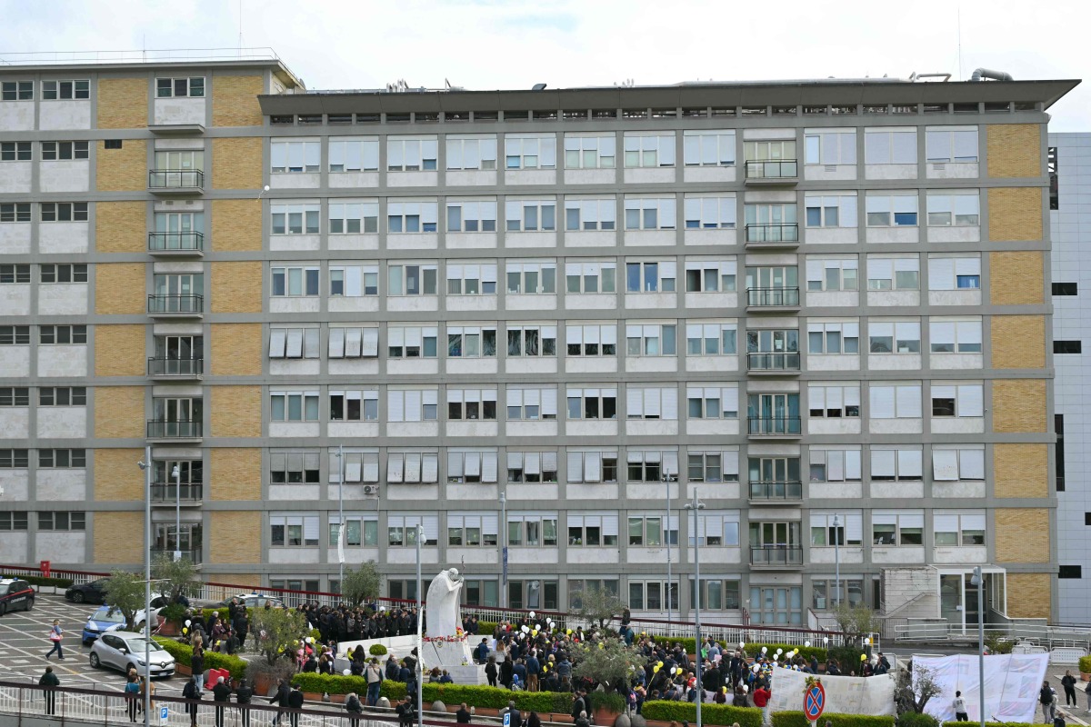 A general view shows children holding white and yellow balloons gathering by the statue of John Paul II outside the Gemelli University Hospital where Pope Francis is hospitalised, in Rome on March 16, 2025. (Photo by Andreas SOLARO / AFP)